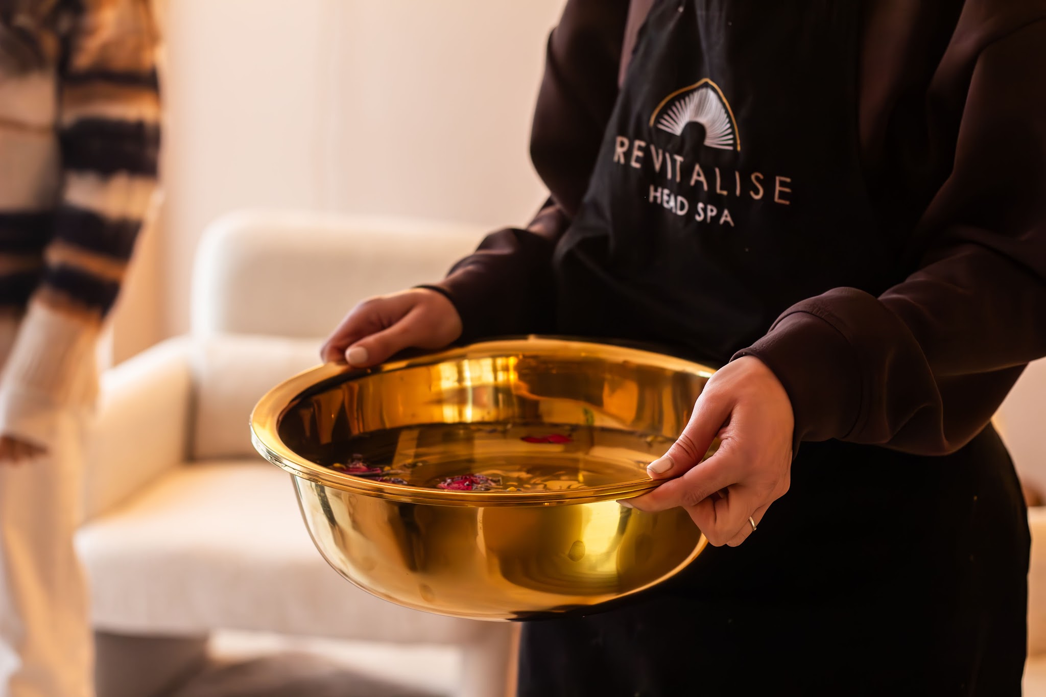 A Revitalise therapist in a black Revitalise Head Spa apron holding a gold foot soak bowl scattered with petals